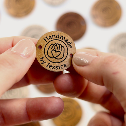 Handmade faux leather tag with 'Handmade by Jessica' text held between fingers against a blurred background of similar badges.