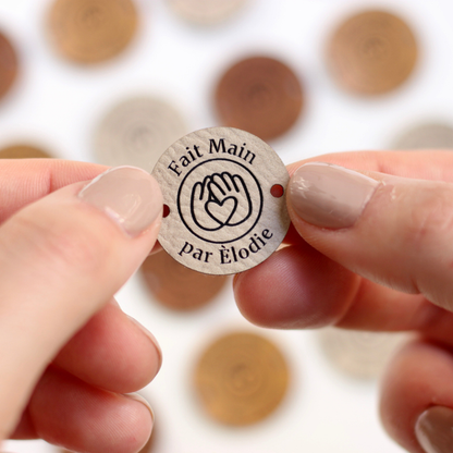 Hand holding a round faux leather label with 'Fait Main par élodie' text against a blurred background of similar labels.