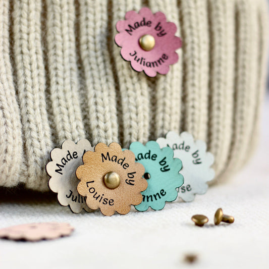 Colorful flower-shaped labels with 'Made by Juhanne' text tags for crochet on a textured knitted surface